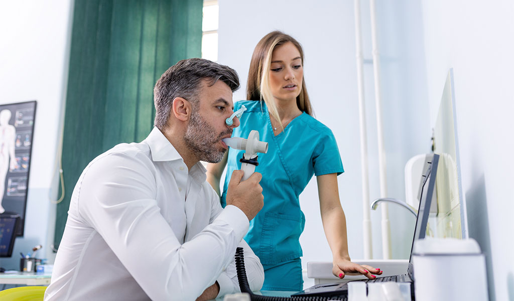 Auf dem Bild sieht man einen Mann mittleren Alters, der gerade einen Lungenfunktionstest (Spirometrie) durchführt. Er hält ein Mundstück und bläst Luft hinein, während eine medizinische Fachkraft, vermutlich eine Krankenschwester oder Ärztin, neben ihm steht und auf einen Computerbildschirm schaut. Sie überwacht offensichtlich den Test und überprüft die Ergebnisse. Der Mann trägt ein weißes Hemd, die Fachkraft trägt medizinische Arbeitskleidung in Türkis. Im Hintergrund sind medizinische Geräte und Diagramme sichtbar, die auf eine klinische Umgebung hinweisen.